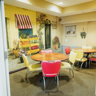 A cozy indoor dining area with round tables and colorful chairs, featuring a retro-style wall mural depicting an outdoor market scene.