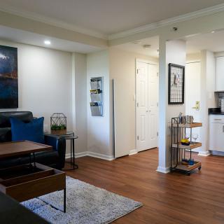 A modern living room with hardwood floors, featuring a cozy couch, coffee table, and a decorative rug. A bar cart is placed near the kitchen entrance.
