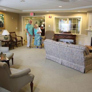 A cozy lounge area with a sofa, chairs, and a piano, where two people are walking through.