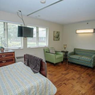 A hospital room with a bed, armchair, and sofa, featuring a wall-mounted television and large windows.