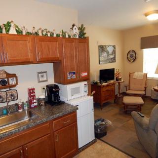 A small, cozy apartment kitchen with a living area in view, featuring wooden cabinets, a microwave, refrigerator, and various decorations.