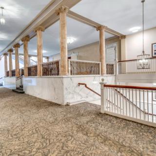 A spacious hallway with elegant columns and carpeted flooring, featuring a staircase with wooden railings.