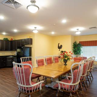 A cozy dining room with a large wooden table surrounded by cushioned chairs and a kitchen in the background.