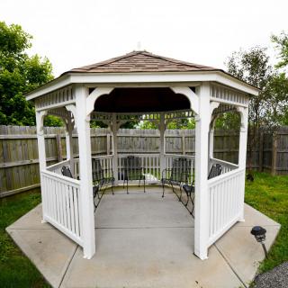 A white wooden gazebo in a fenced backyard surrounded by greenery.