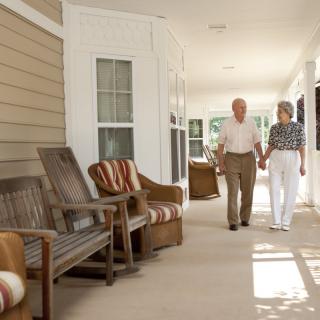 An elderly couple holding hands while walking down a veranda on a sunny day.