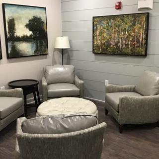 A cozy living room area featuring four gray chairs, a round ottoman, a small side table, a standing lamp, and two landscape paintings on the walls.