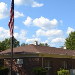 A single-story brick building with an American flag in front, set against a background of blue sky and scattered clouds.