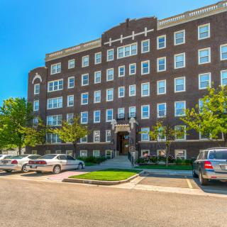 A brick apartment building with multiple floors, surrounded by parked cars and green trees.