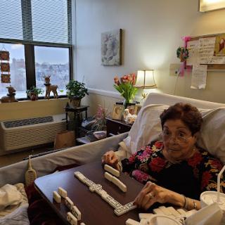 An elderly woman enjoying a game of dominoes in her hospital room, surrounded by plants and personal touches.