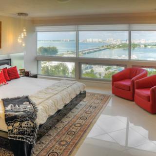 A modern bedroom with large windows showcasing a beautiful lake view, complemented by red chairs and an elegant area rug.