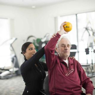 A therapist assists an elderly man with a rehabilitation exercise using a small ball in a clinic setting.
