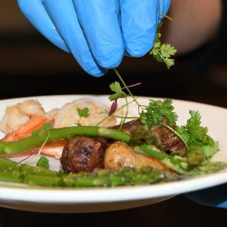 A chef wearing gloves garnishes a dish with fresh herbs and vegetables.
