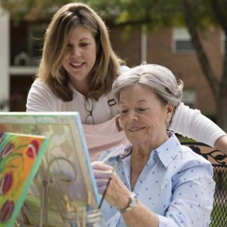 An elderly woman paints on a canvas outdoors, assisted by a middle-aged woman.