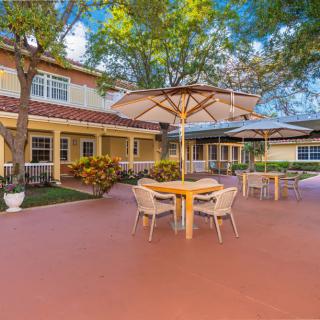A spacious outdoor patio area featuring tables with umbrellas and chairs, adjacent to a building surrounded by trees on a sunny day.