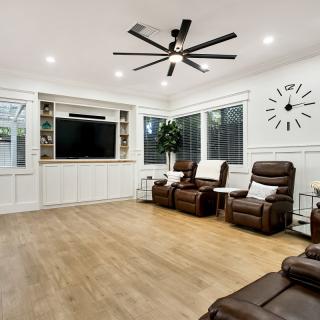 A spacious and modern living room featuring a large TV, recliner chairs, wood flooring, and a ceiling fan.