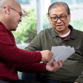 Two people sit together, one showing papers to the other in a comfortable indoor setting.