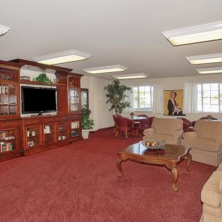 A spacious living room with red carpet, featuring an entertainment center with a TV, comfortable seating, and large windows.