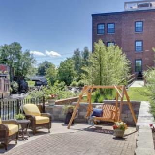 A cozy outdoor patio with a wooden swing and chairs, surrounded by lush greenery and a brick building in the background.