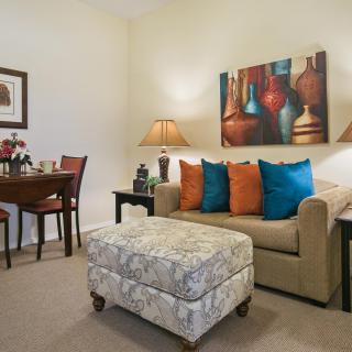 A cozy living room with a sofa adorned with colorful cushions, artwork on the wall, and stylish lamps beside a small dining table.