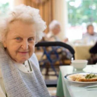 An elderly woman sits at a dining table, enjoying a meal in a comfortable setting.