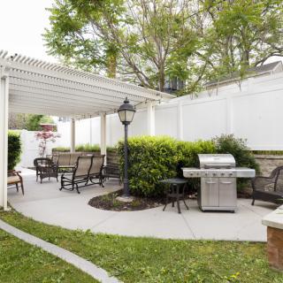 A cozy backyard patio with a pergola, outdoor furniture, and a grill, surrounded by lush greenery and a white fence.