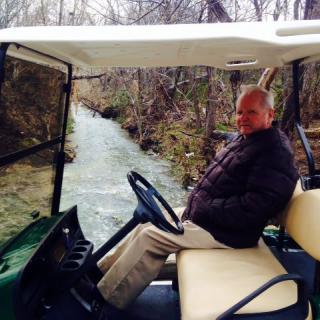 A man sitting in a golf cart near a river with trees surrounding the area.