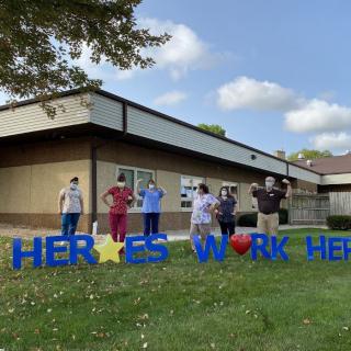 Healthcare workers stand proudly in front of a building with a 'Heroes Work Here' sign, celebrating their dedication.