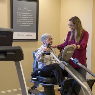An elderly woman receives assistance from a trainer while using exercise equipment in a gym.