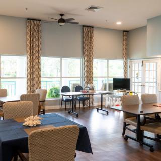A community center room with tables, chairs, large windows, and a computer station.