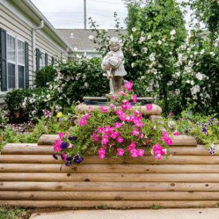 A charming garden scene featuring a small statue atop a wooden planter with vibrant pink flowers cascading down.