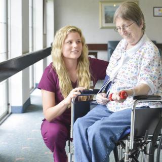 A caregiver kneeling beside an elderly person in a wheelchair, providing assistance.