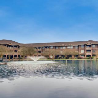 A scenic view of a residential building by a tranquil lake with a fountain under a clear blue sky.