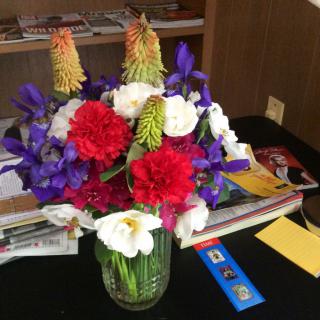 A vibrant bouquet of various flowers in a glass vase, set on a table with magazines and cards.