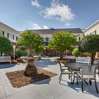 A tranquil courtyard featuring a central fountain, surrounded by trees, benches, and tables with chairs.