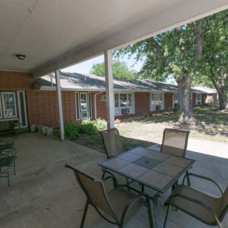 A covered patio area with a table and chairs, adjacent to a brick building and surrounded by greenery.