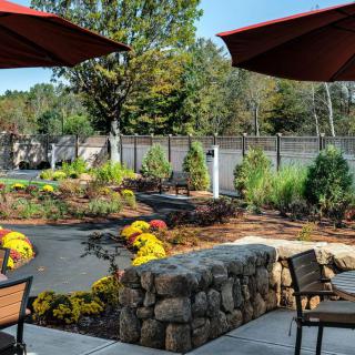 A beautifully landscaped outdoor patio with umbrellas, chairs, and a stone wall surrounded by trees.