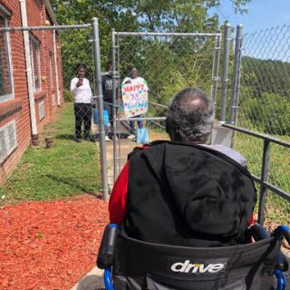 A person in a wheelchair enjoys the outdoors while being assisted by caregivers near a fenced area.