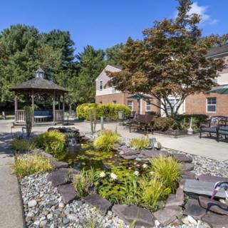 A beautifully landscaped garden featuring a gazebo, a pond surrounded by rocks and greenery, and outdoor seating on a sunny day.