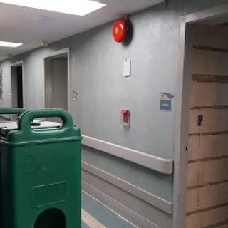 A view of a hospital corridor featuring an emergency alarm and a green trash can.