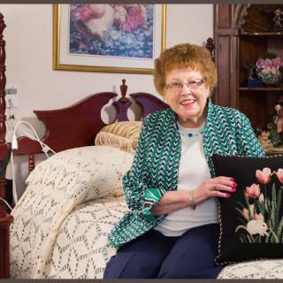 An elderly woman is seated on a bed in a warmly decorated room, smiling and holding a pillow with floral designs.