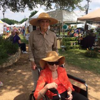 Two people at an outdoor event, one in a wheelchair, both wearing hats.