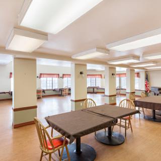 A spacious community room featuring several tables and chairs, illuminated by natural light from the large windows.