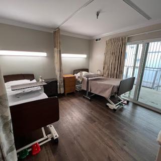 A spacious hospital patient room featuring two adjustable beds, soft lighting, and a view through the window.