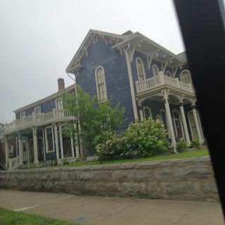 A beautifully crafted historic house with blue walls and elegant porch detailing, surrounded by lush greenery.