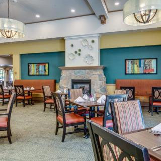 A cozy restaurant dining room with elegant interiors, featuring tables and chairs arranged around a central fireplace and colorful artwork on the walls.
