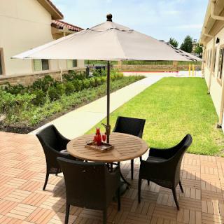 A cozy outdoor seating area featuring a round table with chairs under a white umbrella, surrounded by a lush green garden.