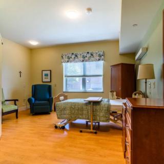 A well-furnished nursing home room with a bed, chair, and window.
