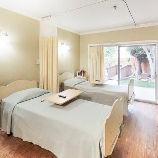 A hospital room with two patient beds, separated by curtains, and a large window providing natural light.