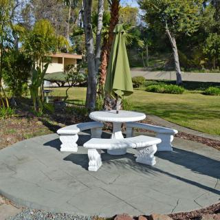 A scenic garden area featuring a white stone table with benches and a green umbrella, surrounded by trees and lush greenery.