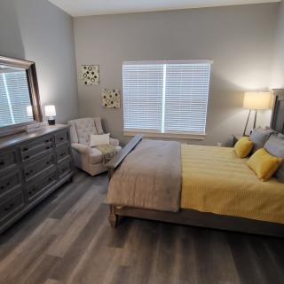 A modern bedroom with a bed, dresser, and armchair, featuring yellow accents and natural lighting.